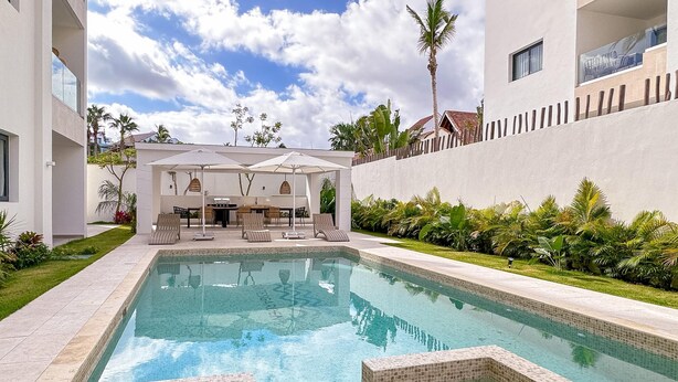 A swimming pool with tiled edges, surrounded by a grassy area and a white wall. There are lounge chairs and umbrellas under a shaded area.