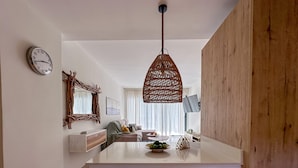 A modern kitchen with a central island, a hanging wicker light fixture, a clock on the wall, and a large window with sheer curtains.