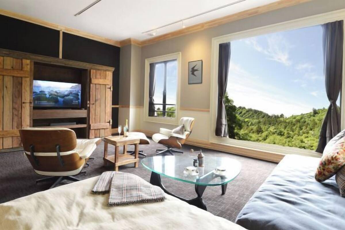 A modern living room with a television, comfortable seating, a glass coffee table, and a large window with a view.