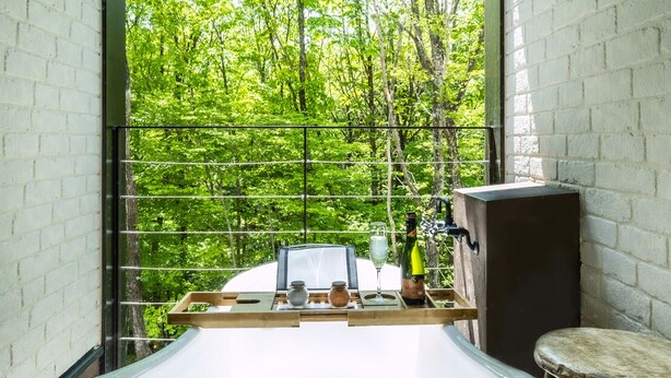 Un bagno moderno con una vasca freestanding, uno sgabello in legno e vista su una rigogliosa foresta verde attraverso un'ampia finestra.