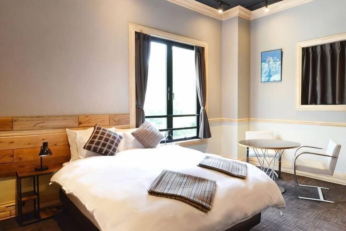 A hotel room with a large bed, a small table, a chair, and a window with curtains.