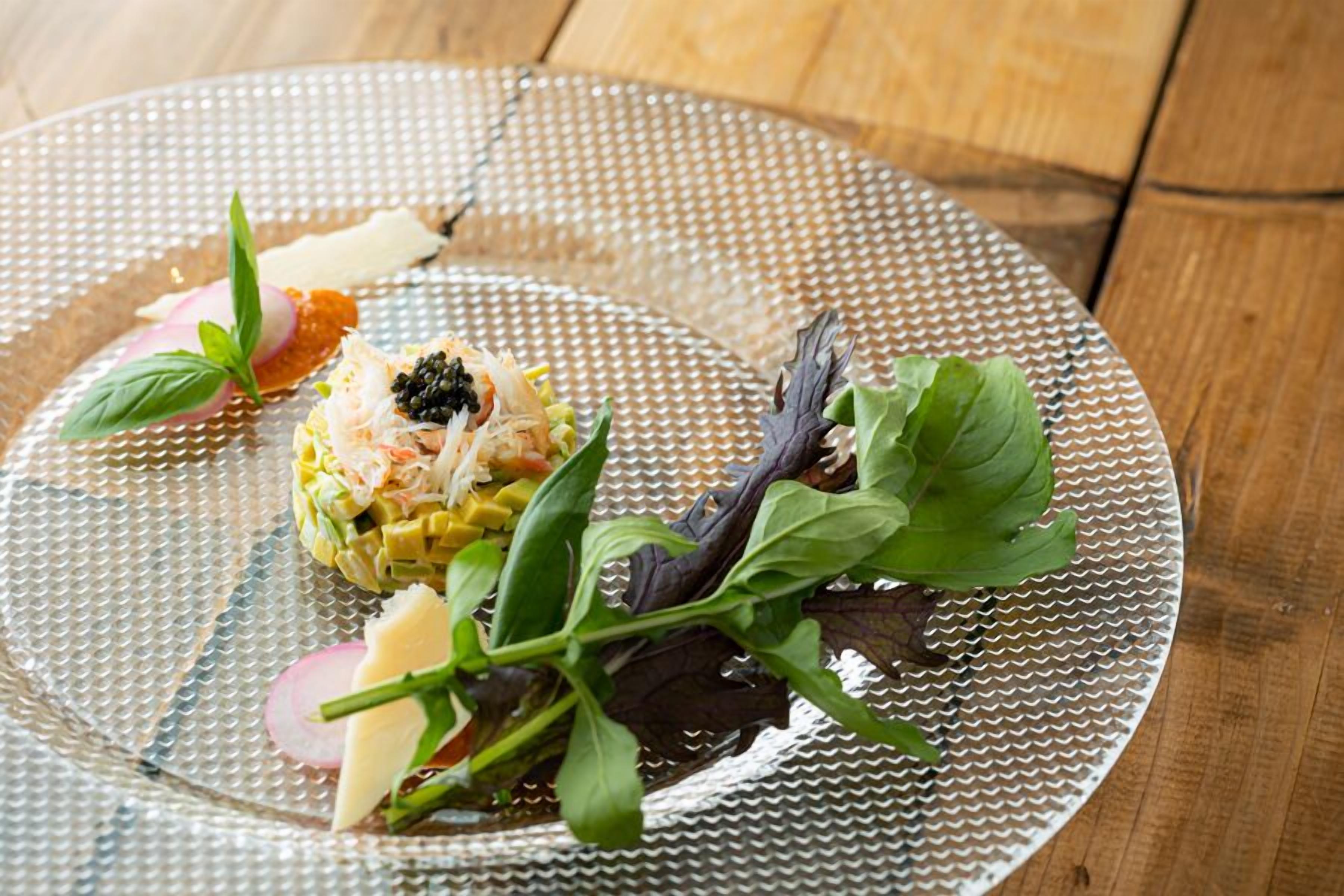 A clear glass plate with a salad, caviar, and greens on a wooden table.