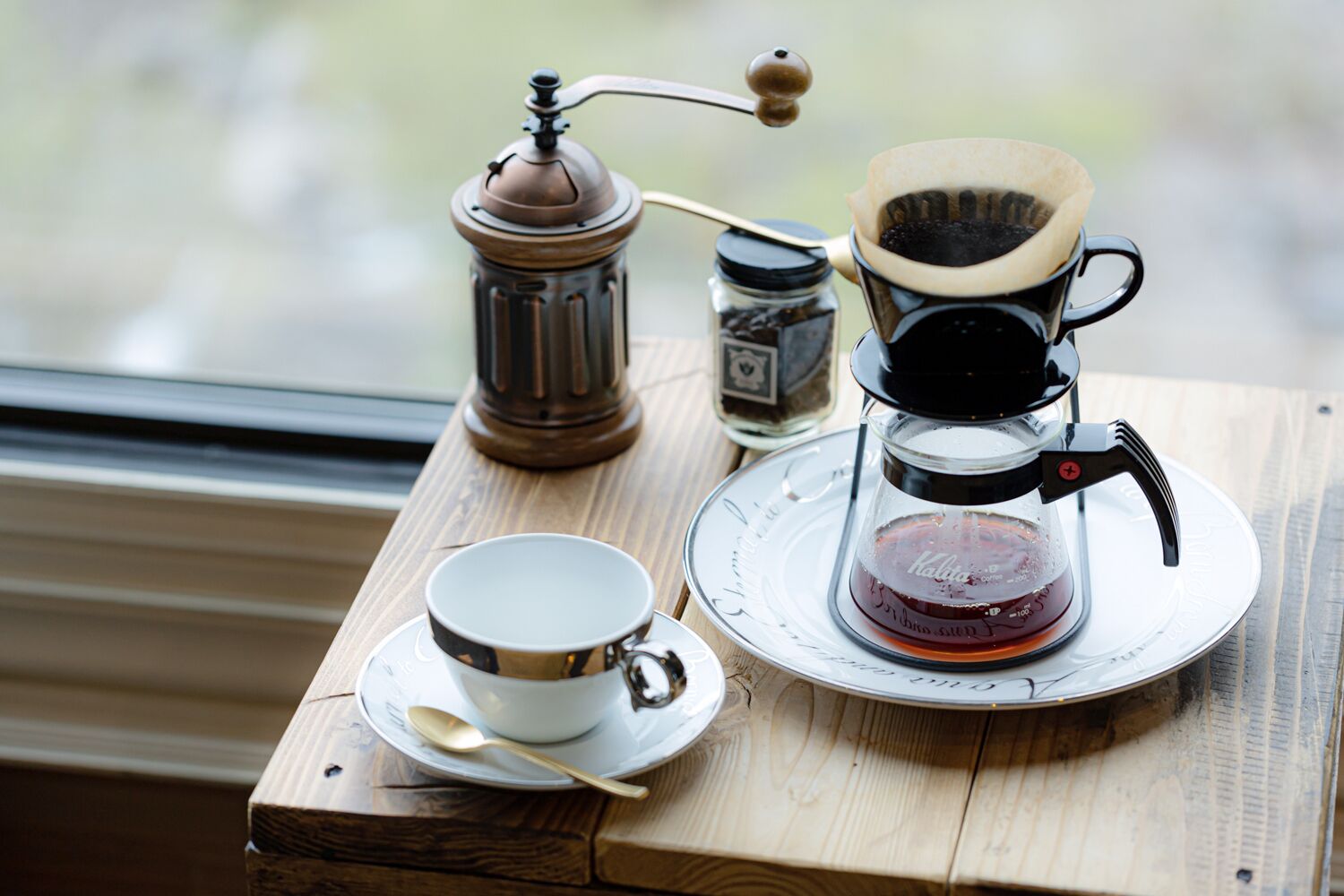 Une machine à café, une tasse et un bocal sur une table en bois près d’une fenêtre.