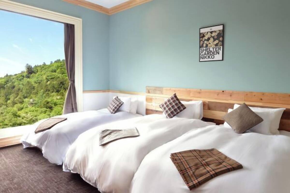 A hotel room with two beds, a large window with a view of greenery, and a framed picture on the wall.