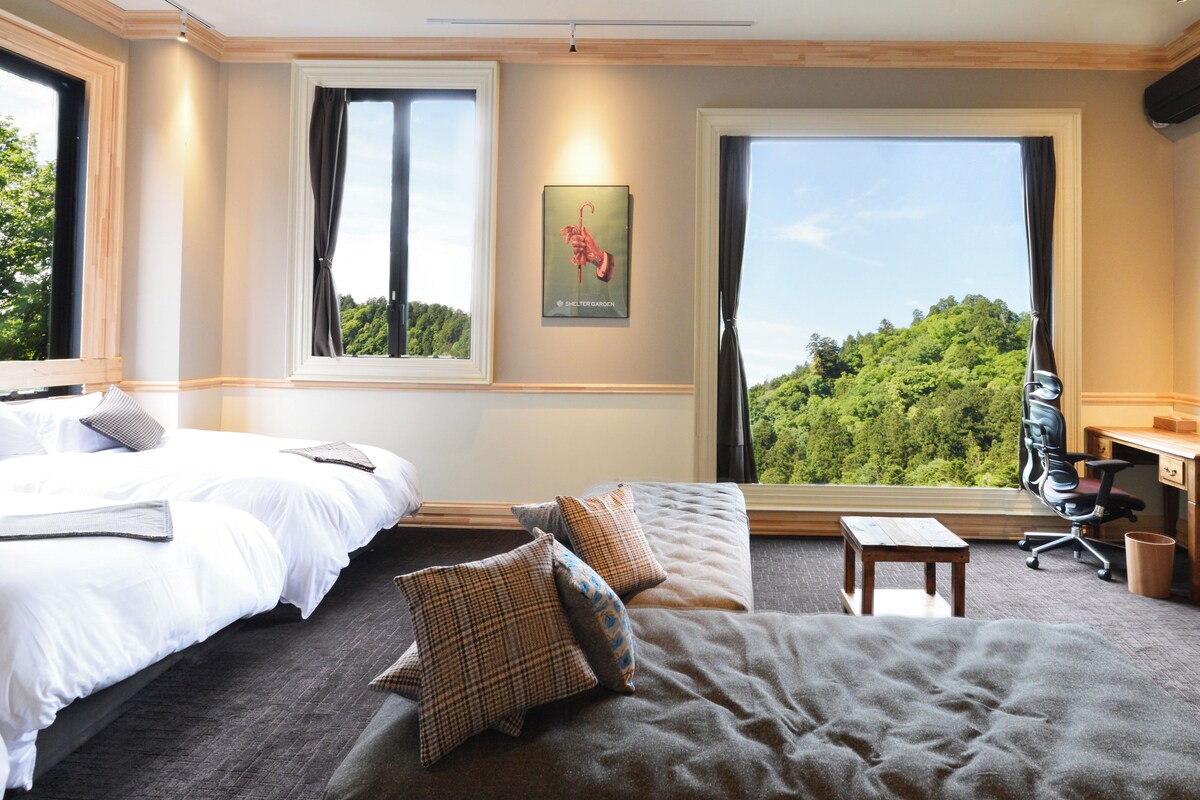 A hotel room with two beds, a sofa, a desk, and a large window offering a forest view.