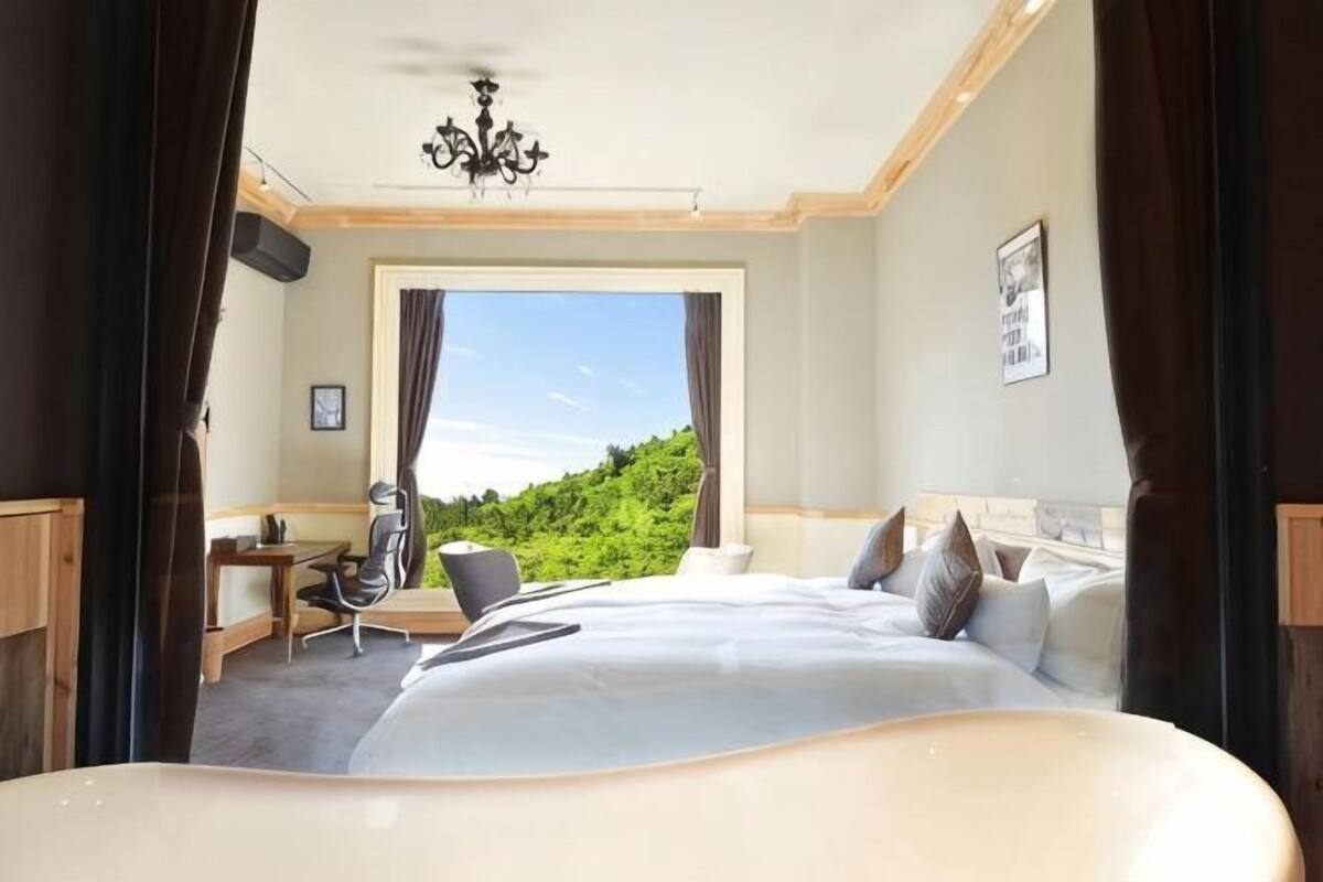 A bedroom with a large bed, a desk, and a chair, with a view of greenery and a mountain.