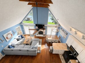 A spacious living room with a high ceiling, a ceiling fan, a fireplace, and a dining area with a wooden table and chairs.