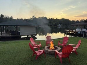 Ein Lagerfeuer mit roten Stühlen davor, mit Blick auf einen ruhigen See und angestellte Boote.