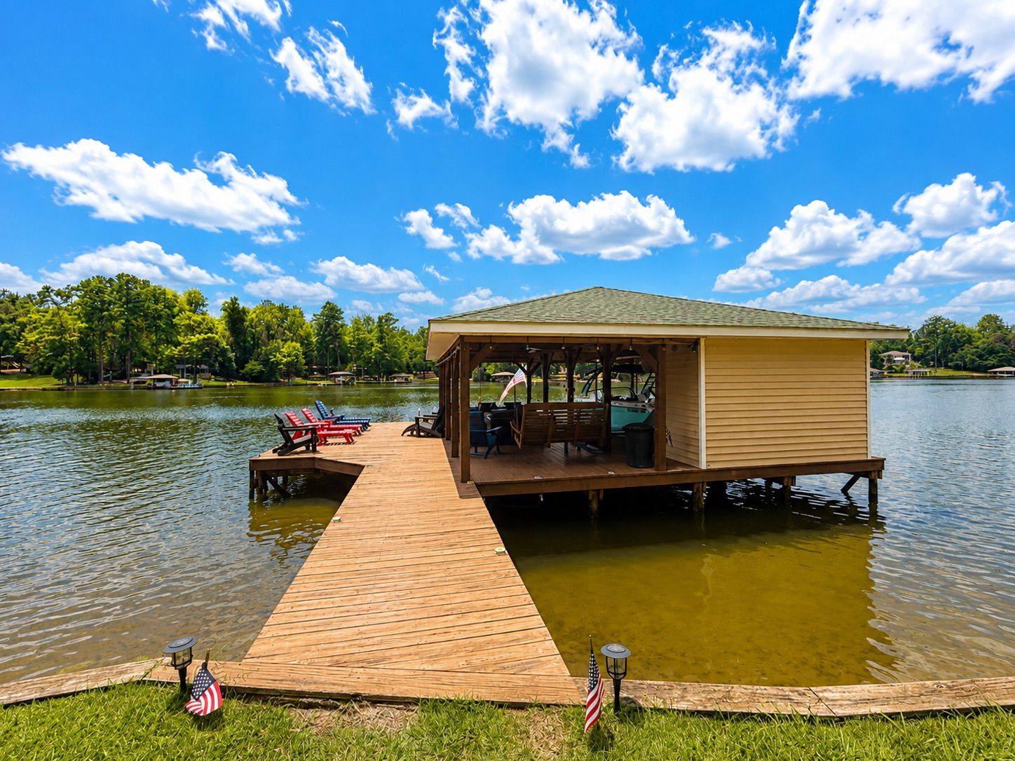 A wooden dock leading to a covered pavilion with seating and a table, overlooking a lake with boats and trees.