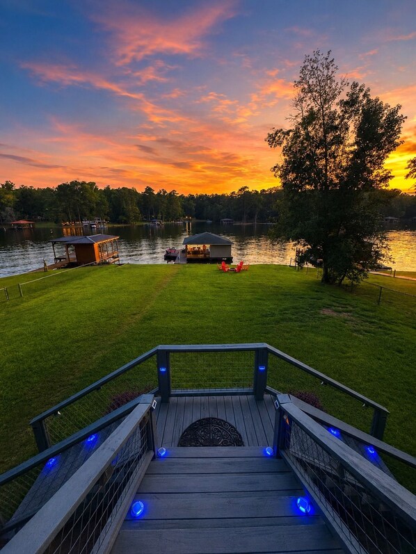 A wooden deck with blue lights, a grassy area, and a lake with boats and a dock.