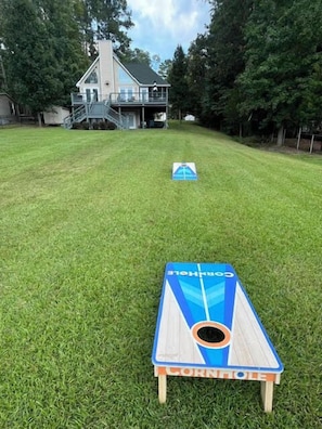 A backyard with a cornhole game set up on a grassy lawn, a house in the background, and trees surrounding the area. - Lake Sinclair Escape - boathouse, 4 bedroom (Sparta)