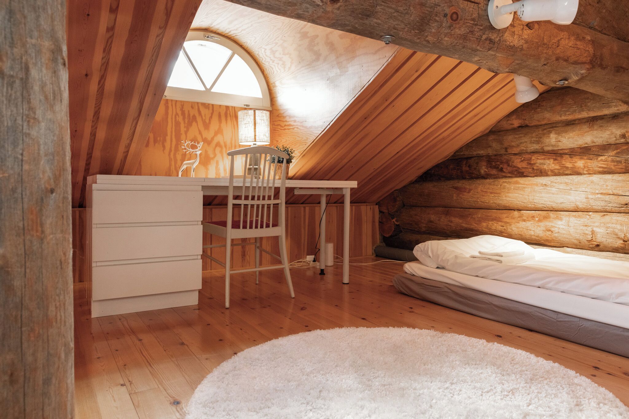 A cozy attic bedroom with a wooden ceiling, a desk with a chair, a bed, and a large window.