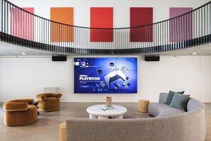 Un área de descanso moderna con una pantalla grande que muestra un anuncio de un juego, asientos cómodos y una escalera con barandilla.