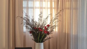 A dining area with a table and chairs, a vase with red flowers, and a window with sheer curtains.