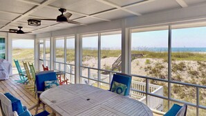 Un espacio al aire libre techado con una mesa y sillas, ventanas amplias con vista a la playa y ventiladores de techo.