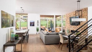 A modern living room with a grey sofa, dining area with a table and chairs, a staircase, and large windows offering a view of a garden. - Luxury Townhome - Exclusive guest access to a members-only golf club and resort amenities. (Sedona)