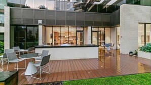 A modern outdoor dining area with a wooden deck, metal chairs, and a glass enclosure. - Garden-View Apartment: Pool, Spa, Gym Access (Caulfield North)