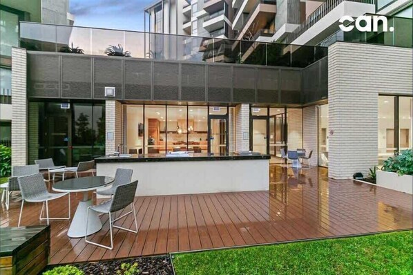 A modern outdoor dining area with a wooden deck, metal chairs, and a glass enclosure.