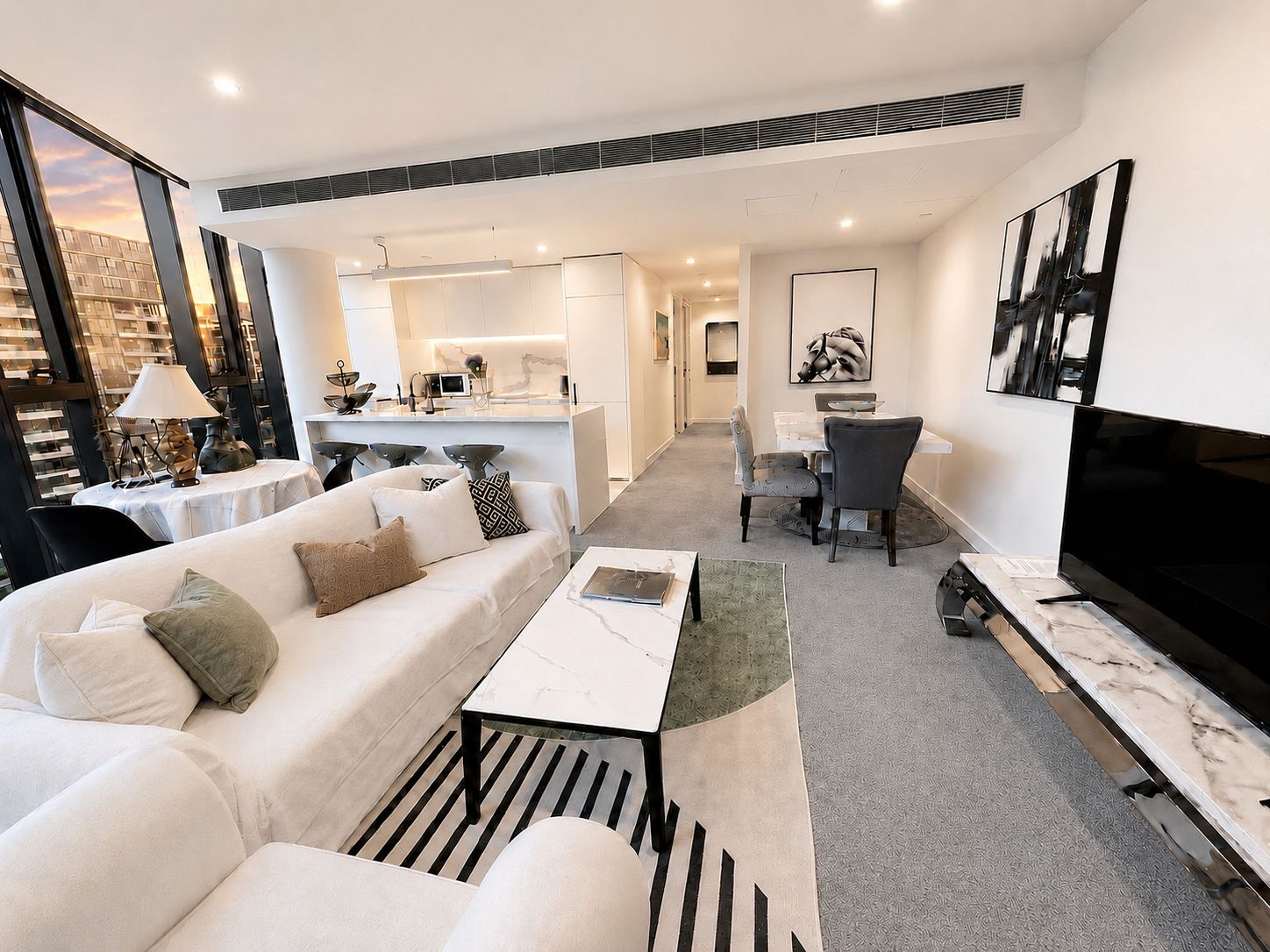 A modern living room with a sofa, coffee table, and a TV. There is a dining area with chairs and a table, and a kitchen in the background.