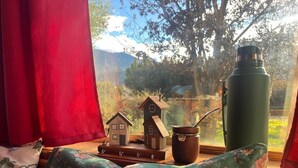 Ein gemütliches Zimmer mit Fensterblick, einem Holz-Hausmodell, einem Thermos und einem blumigen Kissen.