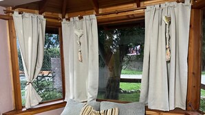 Ein gemütliches Zimmer mit Holzbalkendecke, großen Fenstern mit Vorhängen und einer komfortablen Sitzecke mit Kissen und Decke.