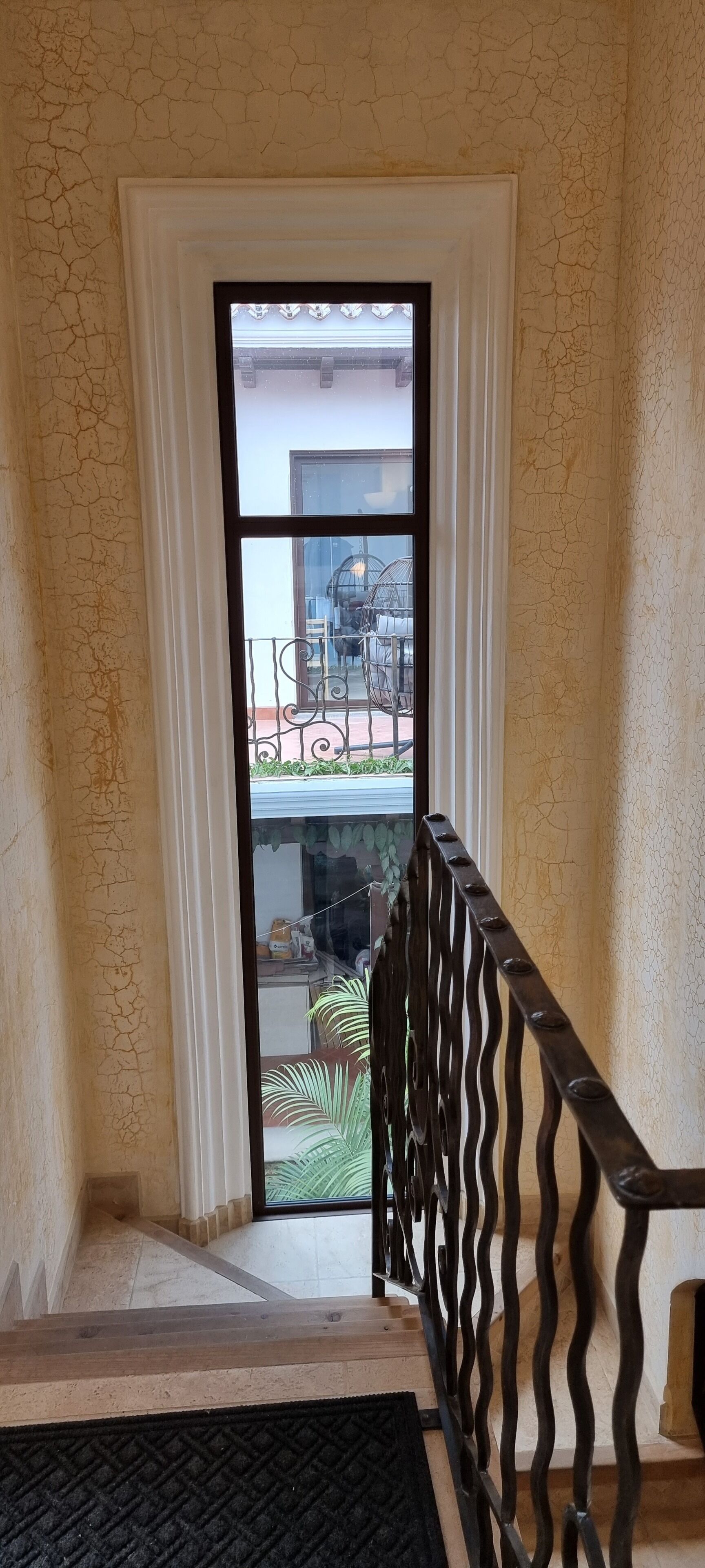 Udsigt fra en balkon med gelænder, en dør med hvidt gardin, og et glimt af havemøbler og planter.