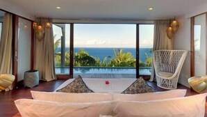 A room with a large window overlooking a scenic view, a white sofa with cushions, a wicker chair, and a small table with a vase.