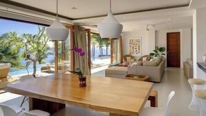 A modern living room with a wooden dining table, white chairs, a sofa, and a view of the ocean.