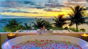 A hot tub filled with water and rose petals, overlooking a beach with palm trees and a sunset.