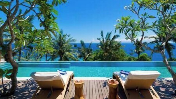 A wooden deck with two lounge chairs and a small table, overlooking a clear blue pool with a view of the ocean and palm trees.