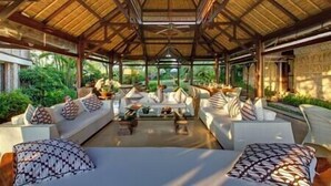 Un vaste espace intérieur doté d’un haut plafond et d’un toit de chaume, comprenant plusieurs coins salon avec des canapés blancs et des coussins à motifs, le tout entouré d’une végétation luxuriante.