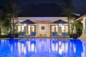 A poolside area with lounge chairs and umbrellas, a covered seating area, and a building with a tiled roof.