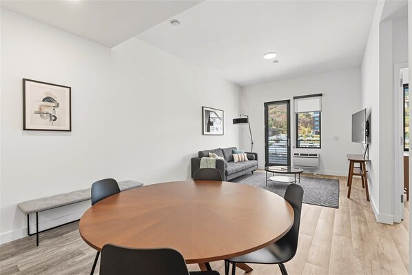 Un salon moderne avec une table ronde en bois, un canapé gris et un banc. Deux tableaux encadrés ornent le mur, ainsi qu’une lampe sur pied et un téléviseur posé sur un meuble.