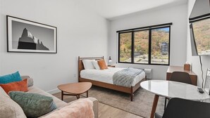 Une chambre moderne avec un lit, une petite table, un canapé et une grande fenêtre donnant sur un paysage de montagne et de arbres.