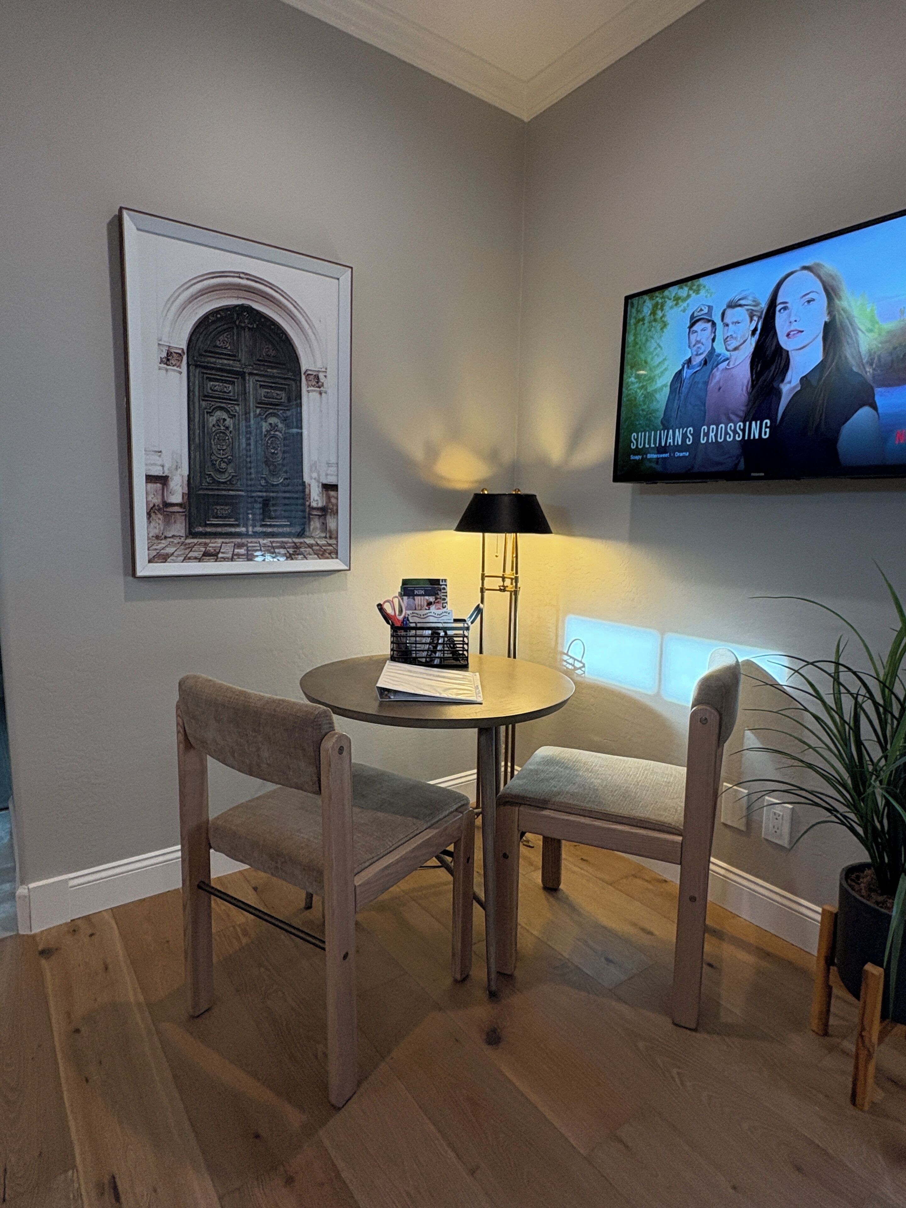Een kamer met een houten vloer, een ronde tafel met twee stoelen, een lamp, een lijst met een foto van een deur, en een televisie waarop 'SULLIVAN'S CROSSING' wordt weergegeven.