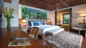 A modern hotel room with a large bed, a wooden desk, and a view of greenery through the window. - Villa Windu Sari by Elite Havens (Seminyak)