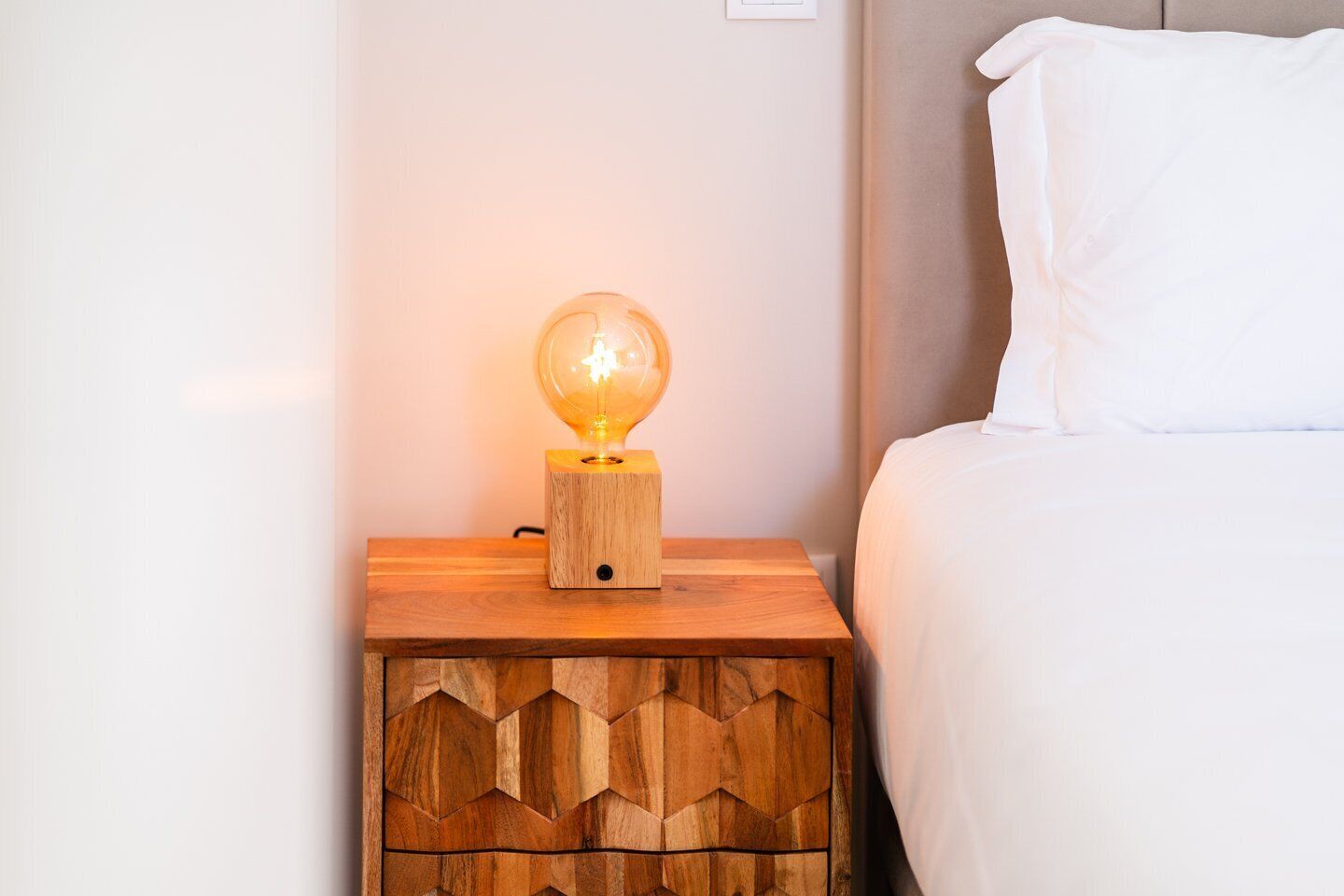Une table de chevet en bois avec une lampe en forme de globe, un lit avec une literie blanche et un mur sobre.