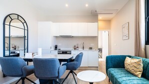 Una cocina moderna con armarios blancos, una zona de comedor con una mesa redonda y dos sillas, y un sofá color turquesa con un cojín estampado.