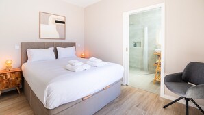 Habitación de hotel moderna con una cama grande, lámparas de noche, una silla y un baño visible a través de una puerta abierta.