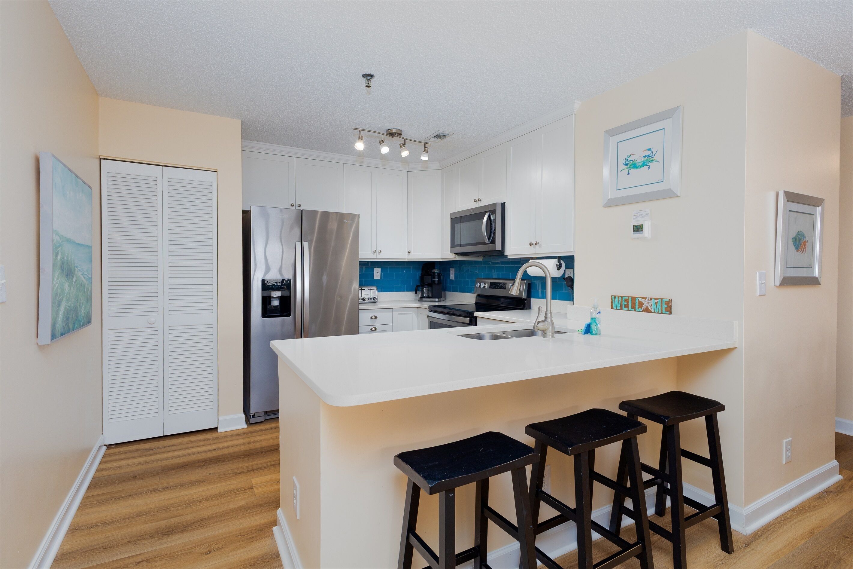A modern kitchen with white cabinets, a stainless steel refrigerator, a microwave, and a sink. There are three stools at the island.