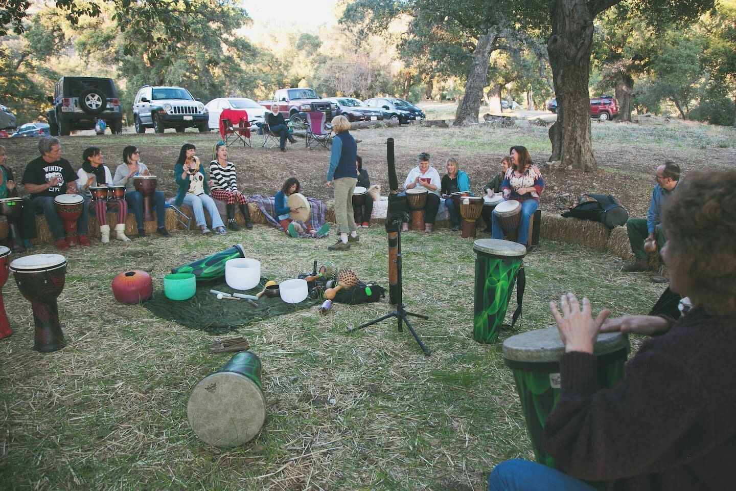 Een groep mensen zit buiten en speelt op drums in een grasveld, met bomen en geparkeerde auto's op de achtergrond.