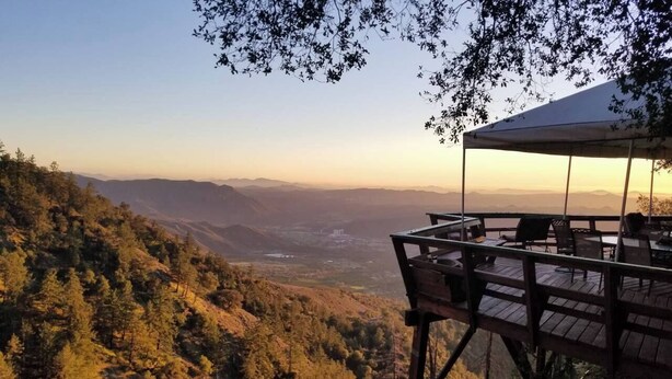A wooden deck with a table and chairs overlooking a mountainous landscape during sunset.