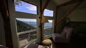 A cozy room with a large window offering a scenic view of mountains and a deck. There is a chair and a small table with a lamp.