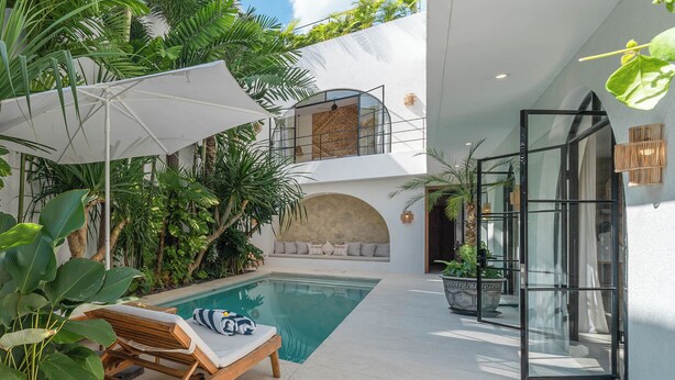 Area piscina interna moderna con una poltrona da relax, un ombrellone bianco e rigogliosa vegetazione tropicale.