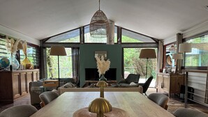 A spacious dining area with a long wooden table, surrounded by comfortable seating. A large window provides ample natural light and a view of greenery outside.