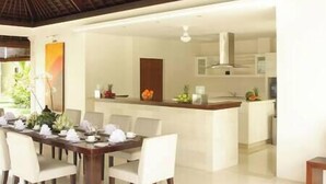 A modern dining area with a central island, white chairs, and a kitchen in the background. - Villa Asante | 4BR Family Villa Near Echo Beach | Pool, Chef (Canggu)