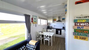 A compact kitchen with white cabinets, a microwave, and a gas stove. There is a dining area with a table and chairs, and a large window with curtains. A motivational sign is visible on the wall. - Chalet Perran Sands, Haven Holiday Park (Perranporth)