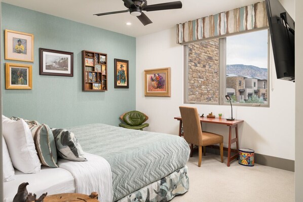 A bedroom with a bed, a desk, and a chair. There are framed pictures on the wall and a window with a view of buildings.