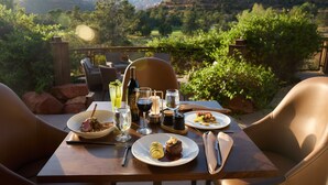 Mesa al aire libre con comida, vasos y una botella, rodeada de sillas, con un paisaje montañoso de fondo.
