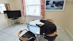 Een kleine, verzorgde kamer met een ronde eettafel, vier stoelen, een televisie en een ingelijste foto aan de muur.